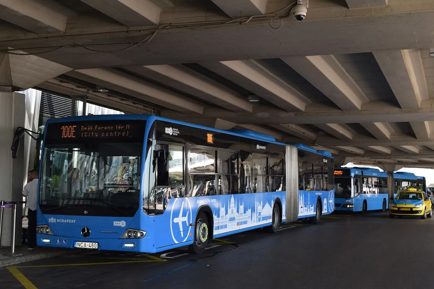 A blue 100E Airport Express bus signed for Deák Ferenc tér, waiting under the terminal canopy