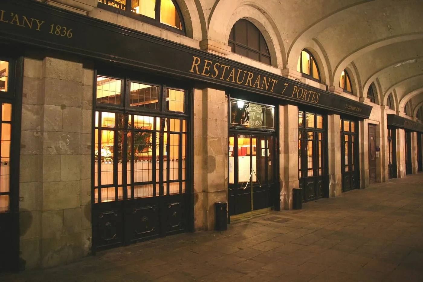The 7 Portes restaurant front under a stone arcade at night, its sign reading Restaurant 7 Portes, with lit windows behind dark wooden frames
