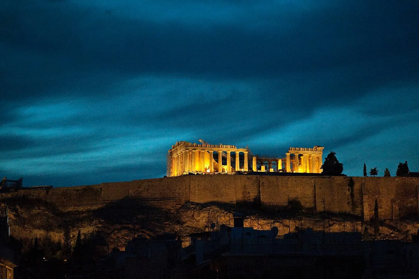 The floodlit Parthenon glowing amber on the Acropolis against a deep blue night sky, with the dark roofs of the city below