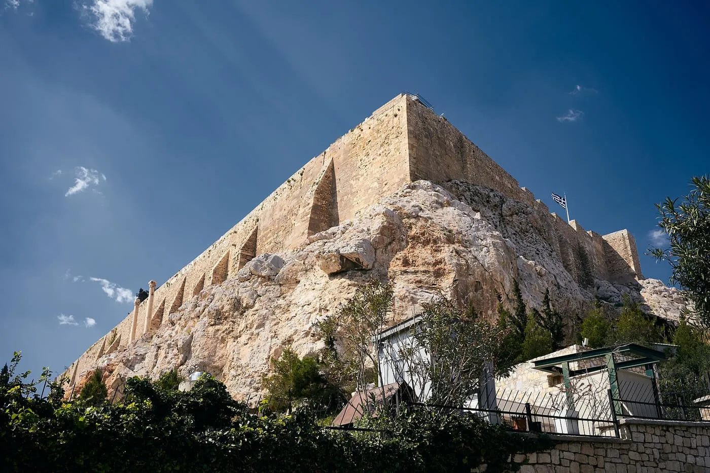 The rock and stone walls of the Acropolis rising steeply against a deep blue sky above the roofs and gardens of Plaka, with a Greek flag on the wall