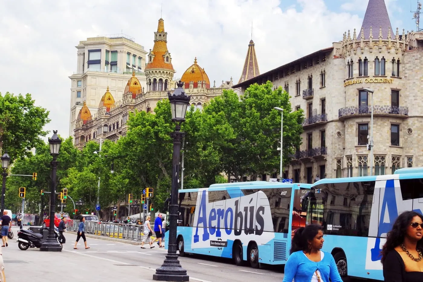Two pale-blue Aerobús coaches parked at Plaça de Catalunya, below the turreted buildings at the foot of Passeig de Gràcia