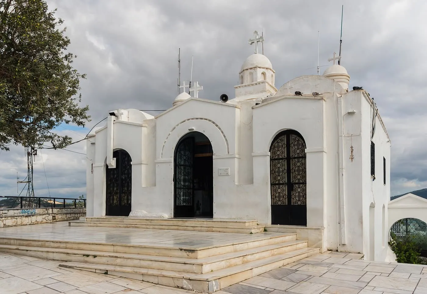 The whitewashed chapel of Agios Georgios on the summit, with three small domes, black iron doors and marble steps under a cloudy sky