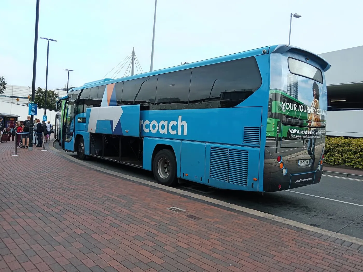 A blue Aircoach coach with its luggage hold open at a kerb at Dublin Airport, passengers queuing beside it