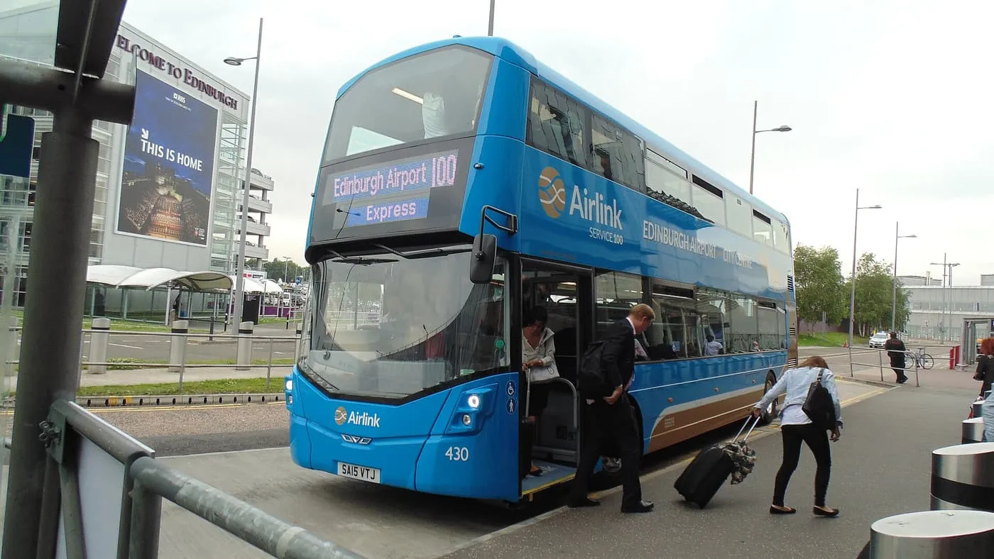A blue double-decker Airlink 100 bus at the airport with passengers boarding and a suitcase being wheeled to the door