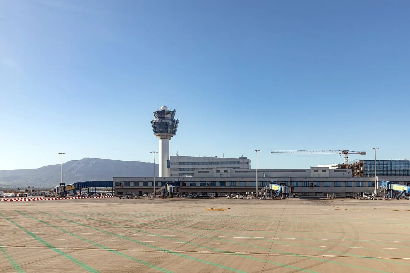 The control tower and low terminal buildings of Athens International Airport seen across the apron, with a hill behind