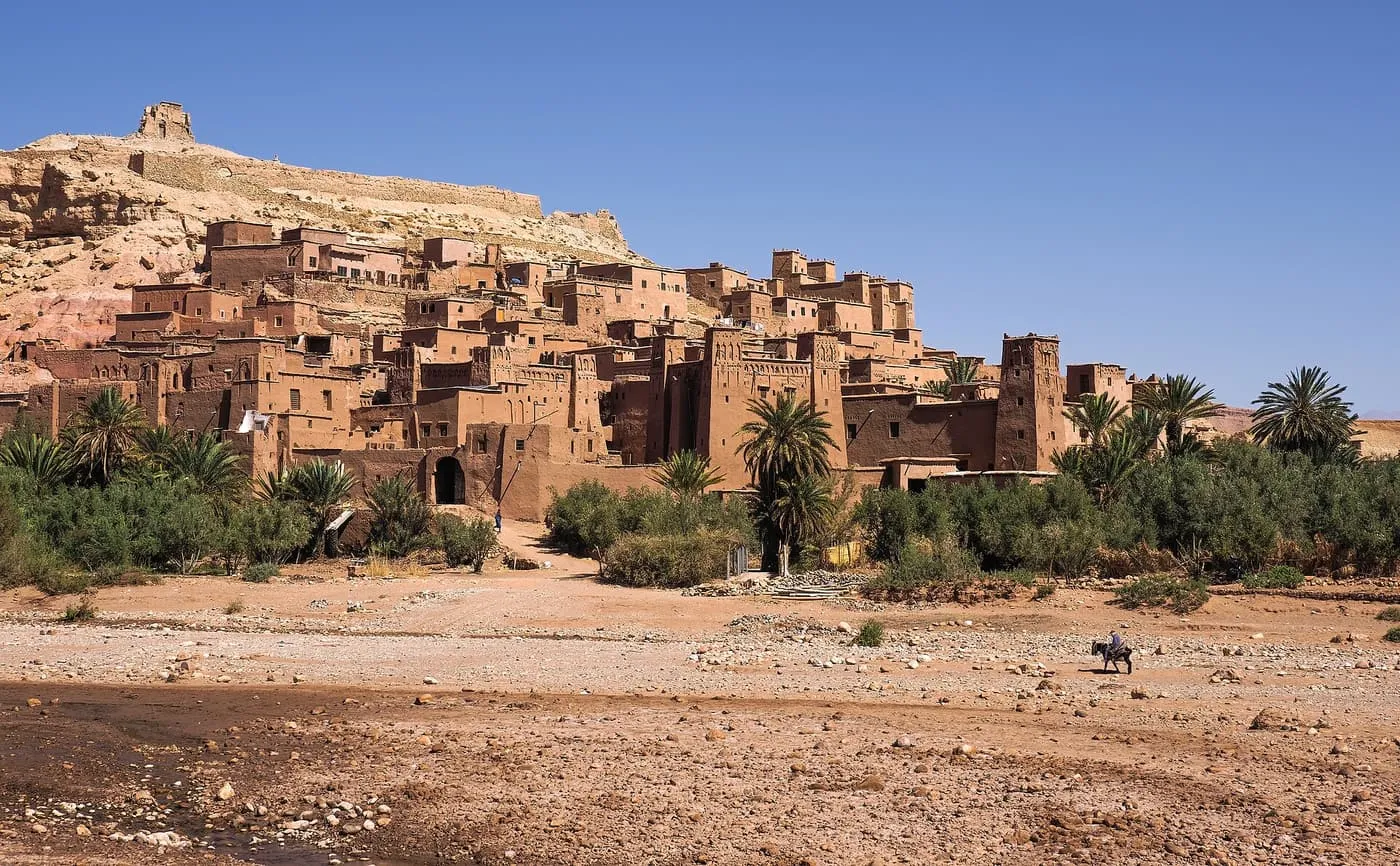 The earthen buildings of Aït Benhaddou rising up a hillside above a dry riverbed and palm trees