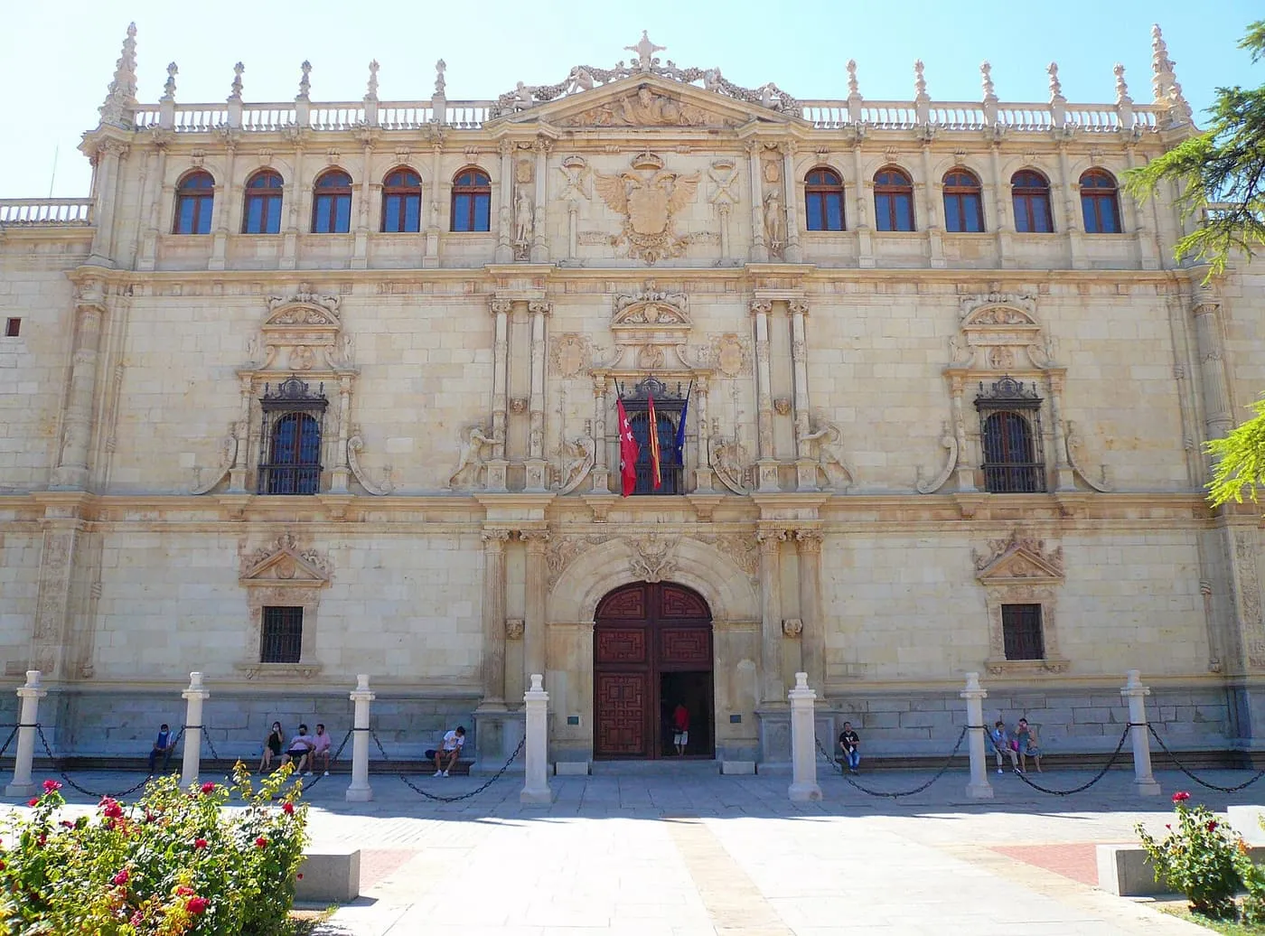 The carved stone façade of the Colegio Mayor de San Ildefonso in Alcalá de Henares, with a heavy wooden door at its centre and rose bushes in front