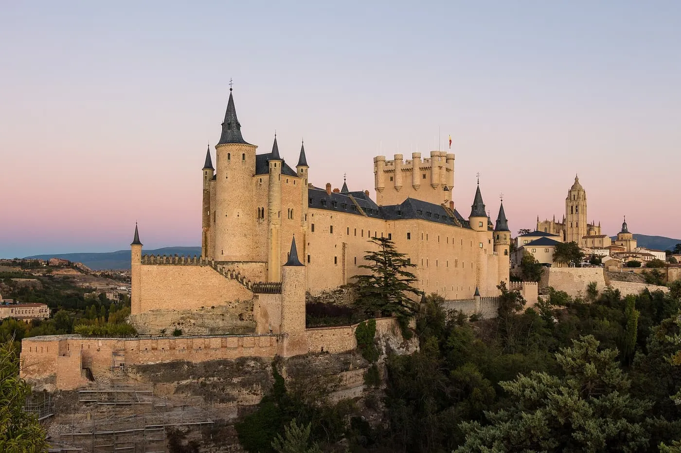 The Alcázar of Segovia on its rock at dusk, with slate-roofed turrets, the square Torre de Juan II and the Cathedral tower behind