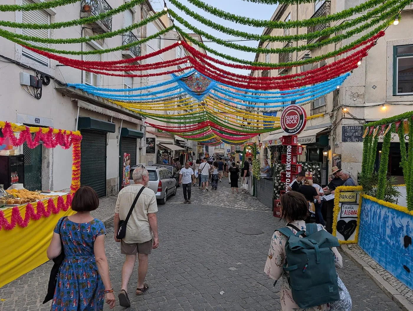 A cobbled Alfama street hung with red, blue, yellow and green paper streamers, with festival stalls and strolling visitors