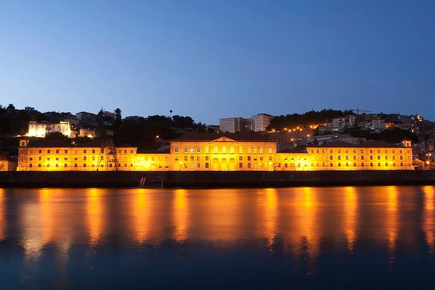 The long lit façade of the Alfândega do Porto at dusk, reflected in the Douro