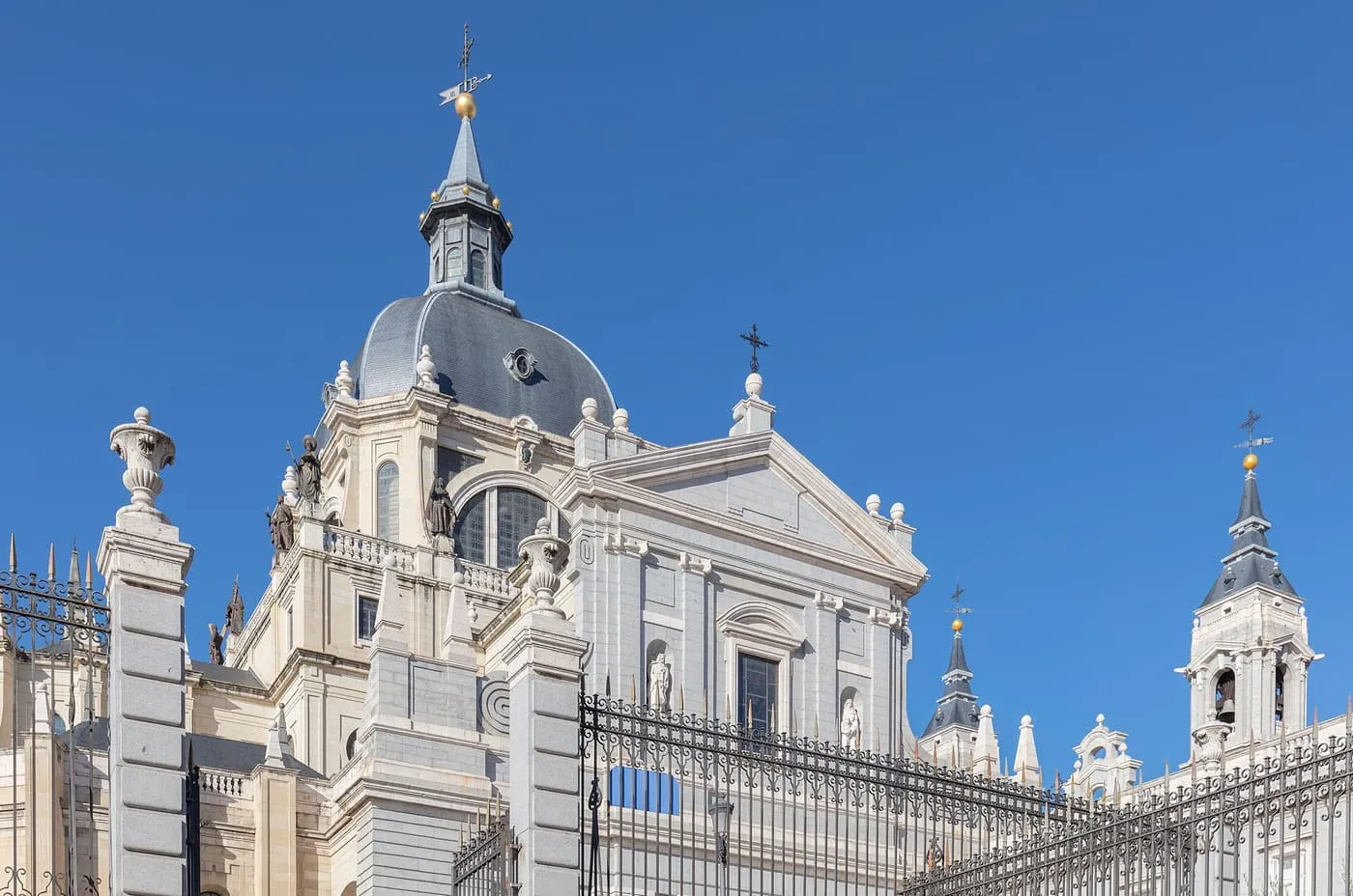 The dome, lantern and bell tower of the Almudena Cathedral in pale stone and grey slate, behind black iron railings under a blue sky