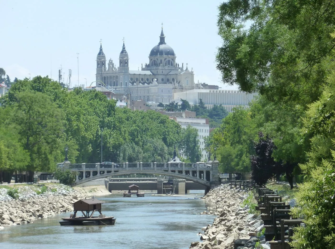 The domed Almudena Cathedral above trees beside the Manzanares, with a stone bridge crossing the river below