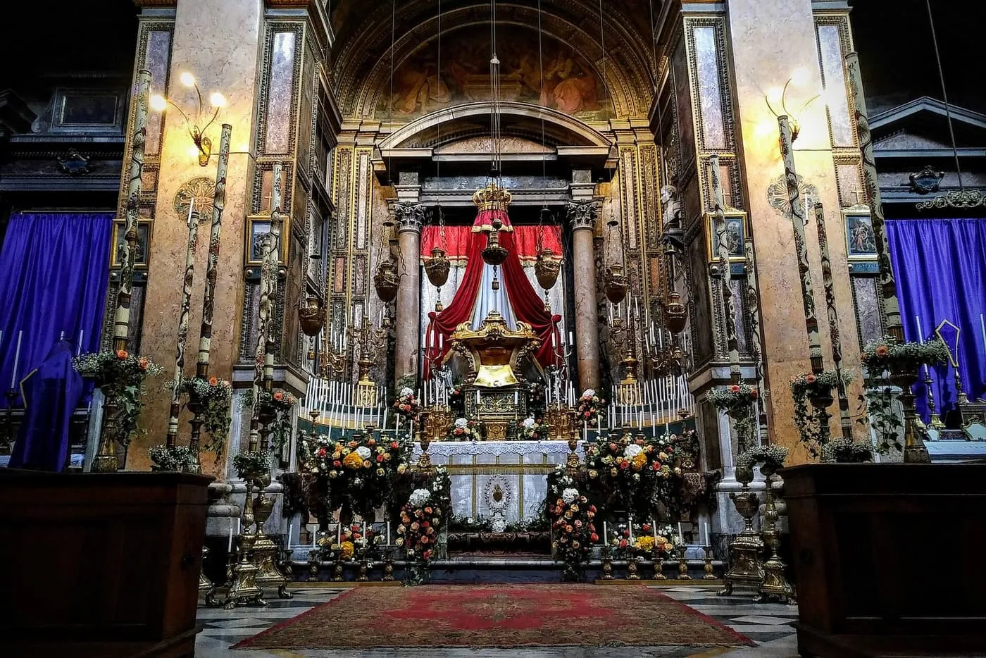 The altar of repose in a Roman church, hung with red drapes and banked with flowers and candles between purple-veiled side altars