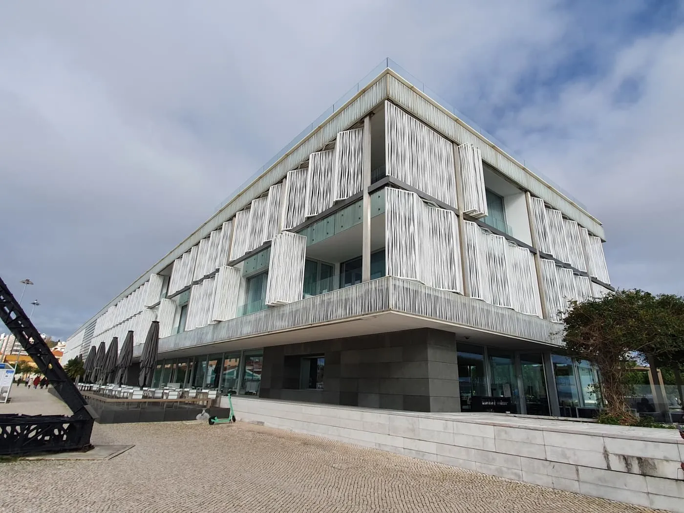 The white-shuttered facade of Altis Belém Hotel & Spa seen from the quay, with its terrace tables and a glass-edged roof