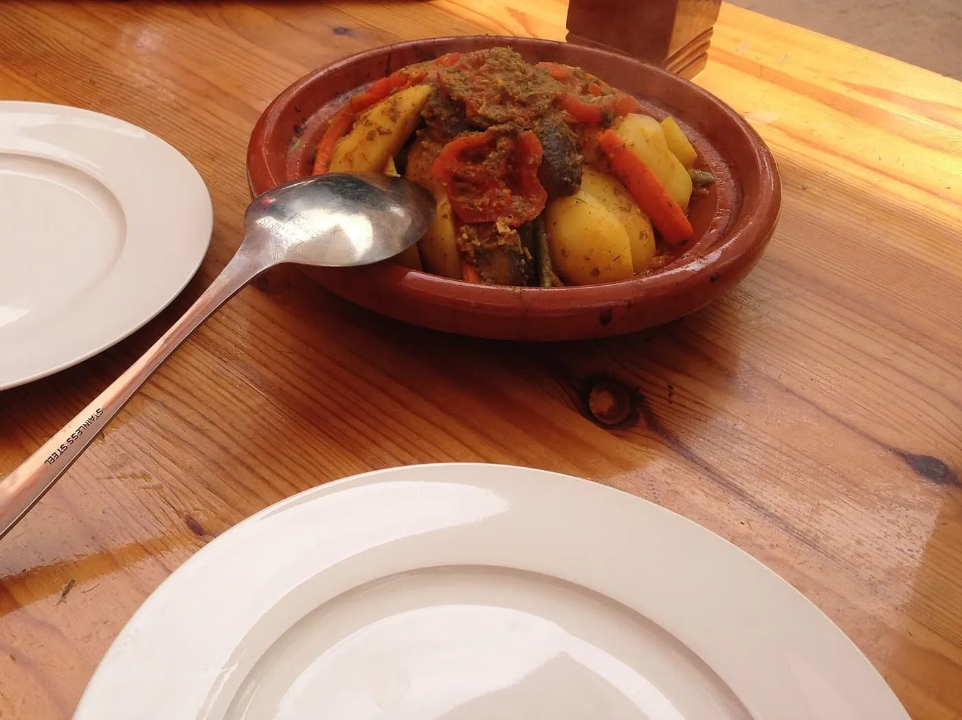 A tajine of lamb, potatoes and carrots in a glazed terracotta dish on a wooden table beside empty plates