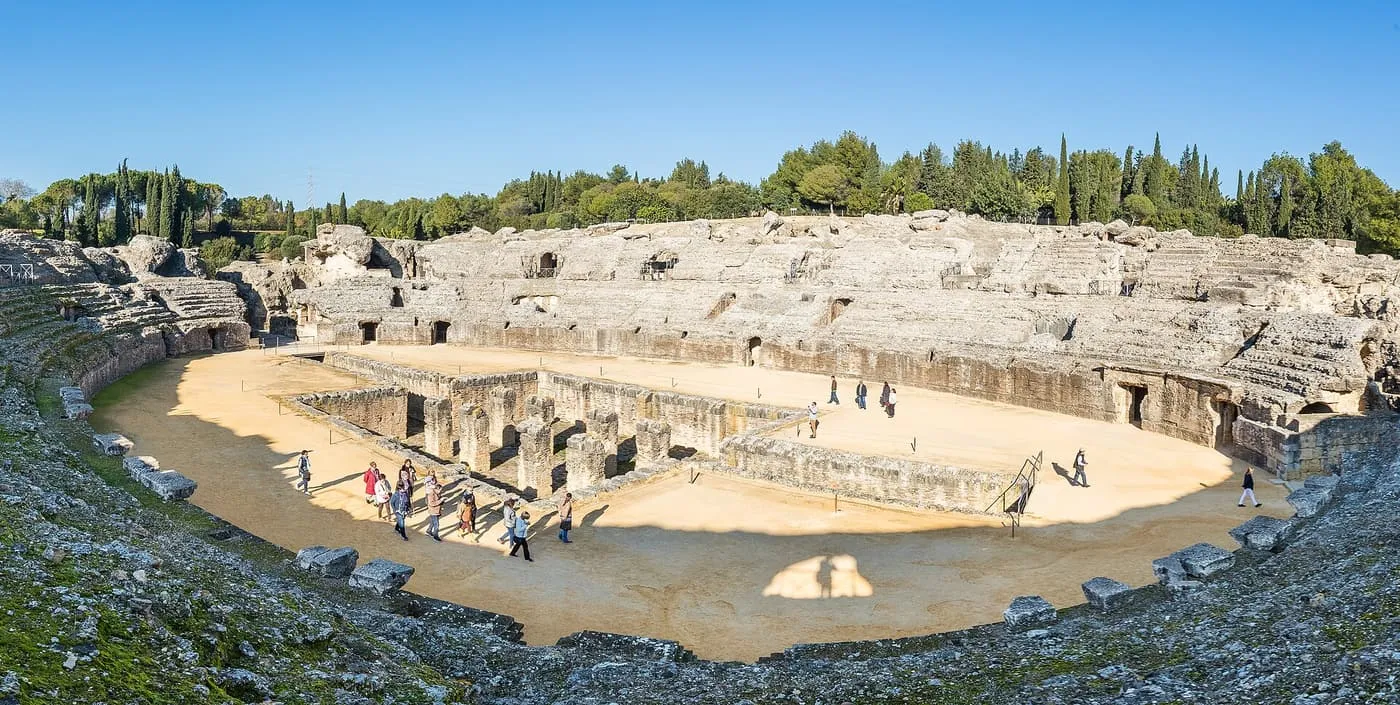 The oval arena of the Roman amphitheatre at Itálica, with the brick substructure at its centre, the weathered tiers of seating and a few visitors walking across the sand
