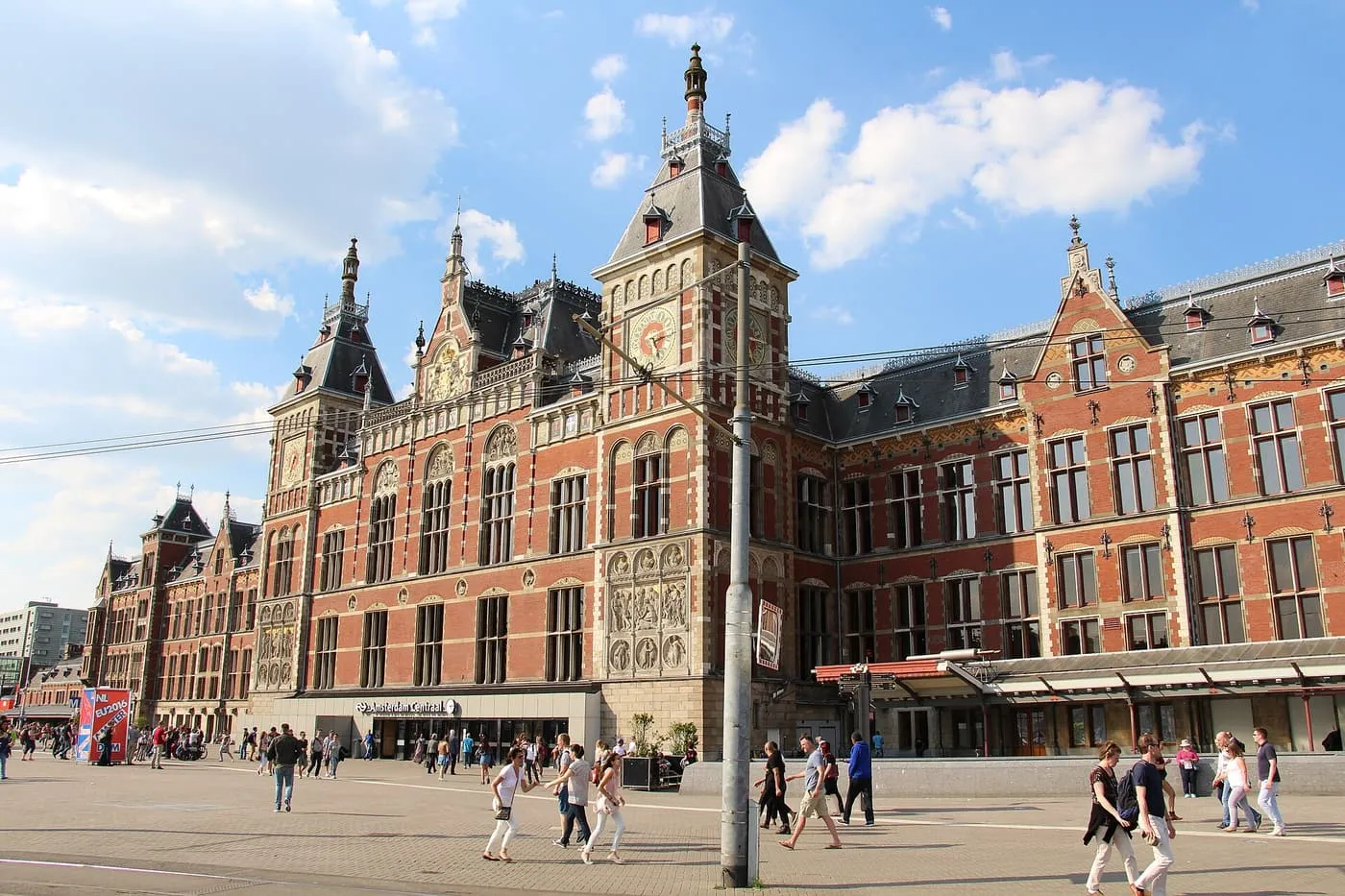Amsterdam Centraal's red brick and stone station building with towers and clock faces, seen across Stationsplein with people crossing the square