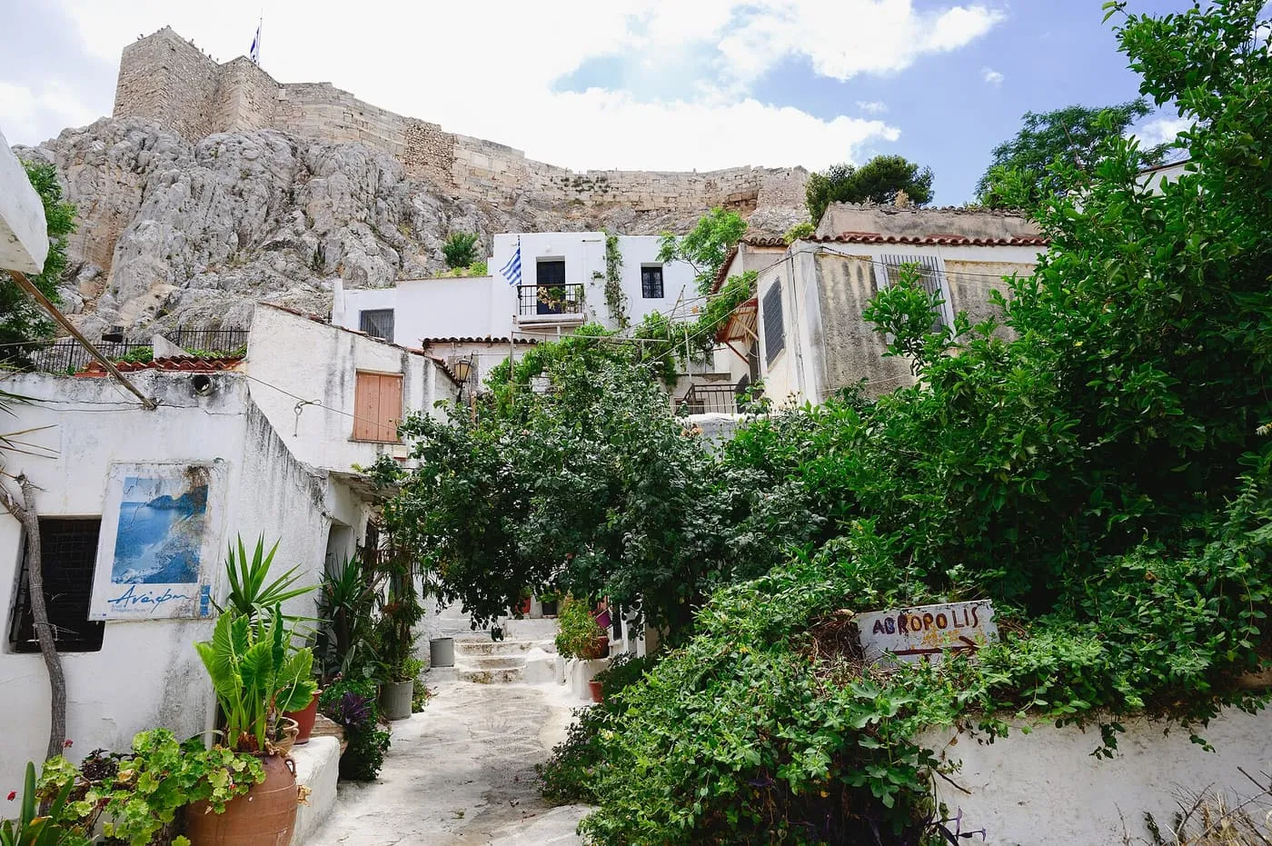 A whitewashed lane in Anafiotika between small houses and green plants, with the rock and walls of the Acropolis rising above