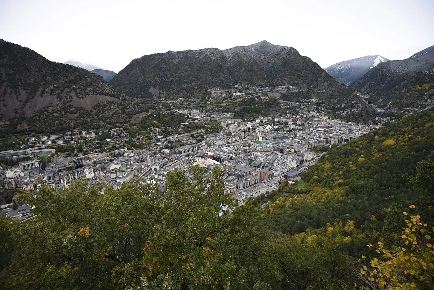 Andorra la Vella packed into a mountain valley, seen from a wooded hillside above, with dark Pyrenean ridges and a dusting of snow behind