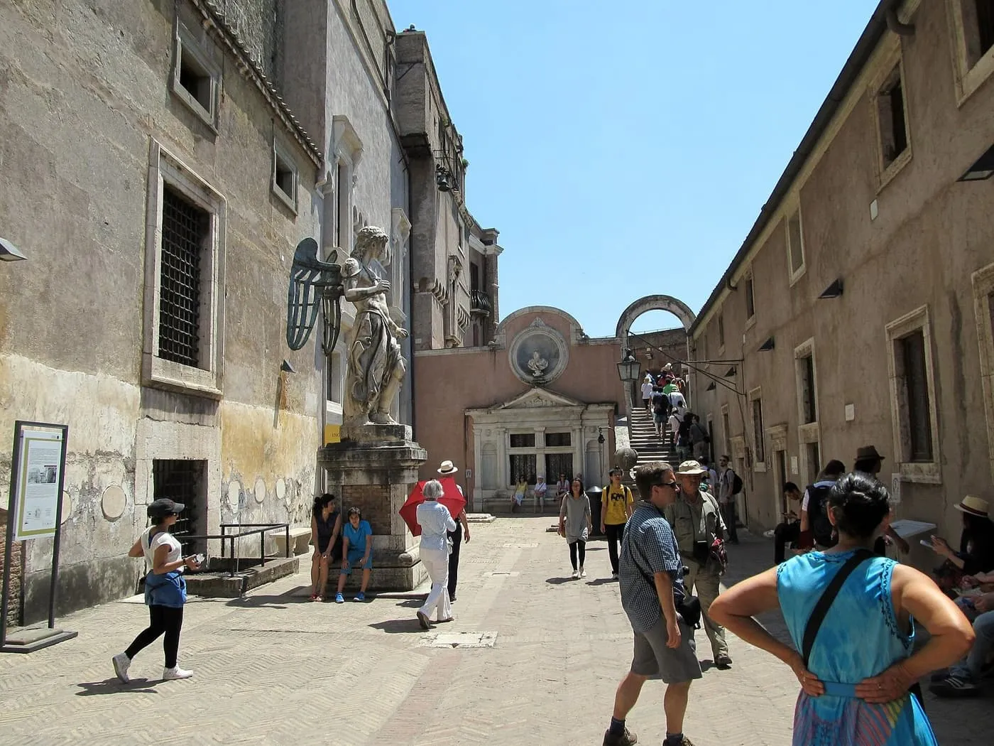 Visitors in the Angel's Courtyard of Castel Sant'Angelo, with the marble angel on its pedestal at the left and a stair climbing at the back