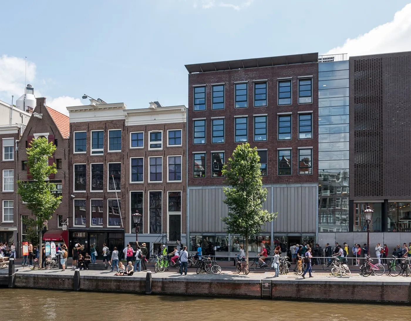 The Anne Frank House on the Prinsengracht, a row of brick canal houses beside the museum's modern brick and glass extension, with people on the quay