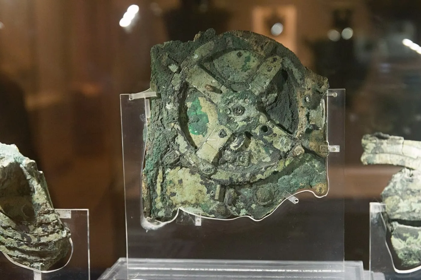 The corroded green bronze main fragment of the Antikythera Mechanism, with gear wheels visible, mounted on clear plastic in a display case