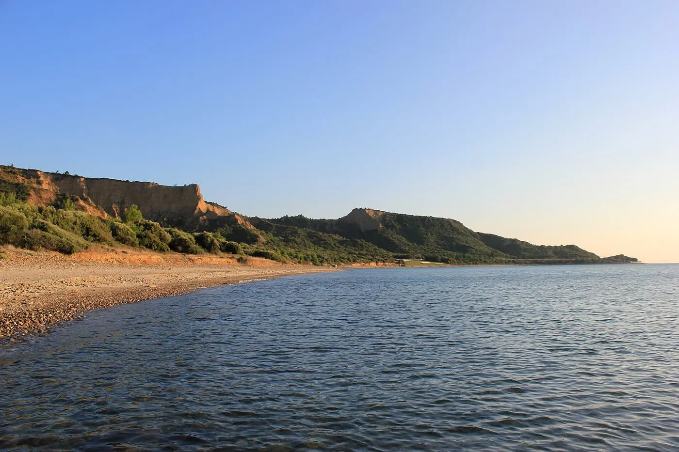 A shingle beach and low scrub-covered cliffs curving along a calm sea at Anzac Cove in evening light