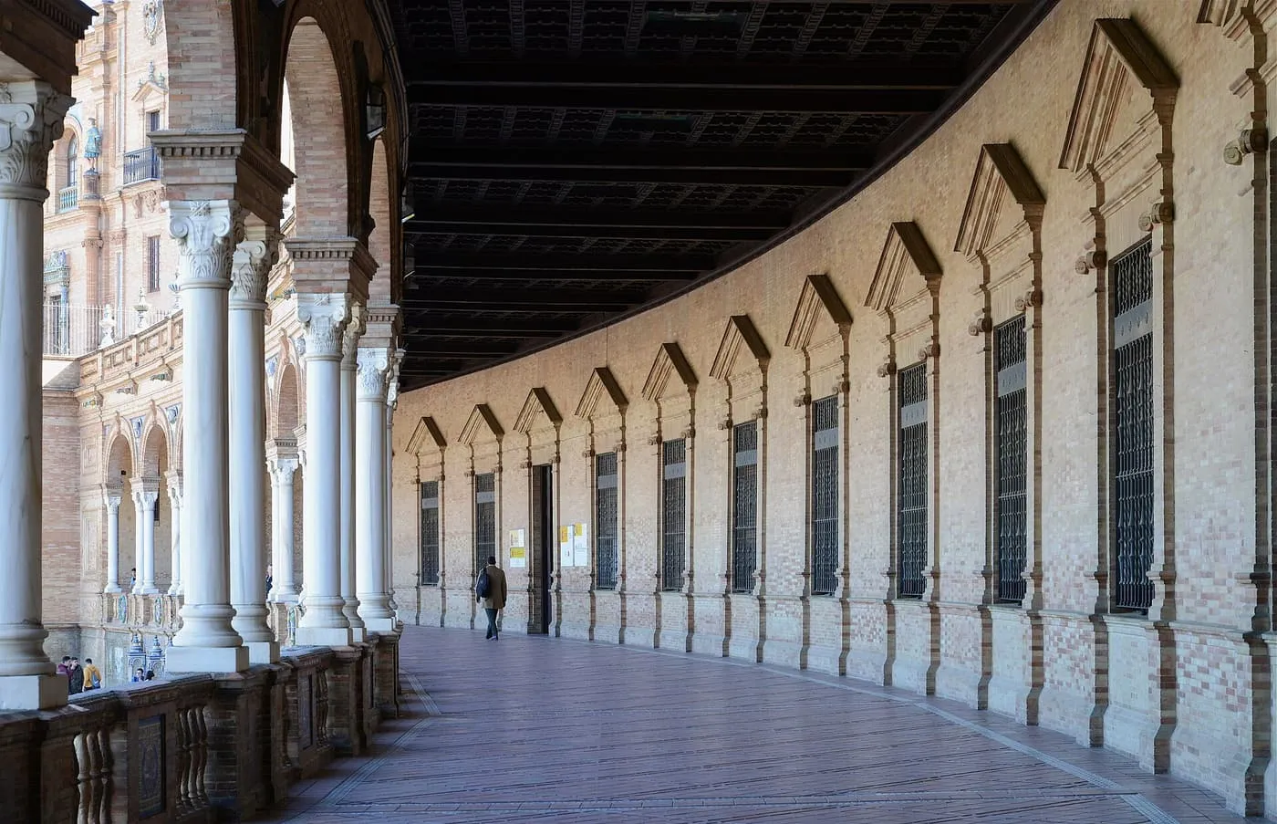 The curved arcaded gallery of the Plaza de España, with white marble columns on the left and a row of barred windows on the right