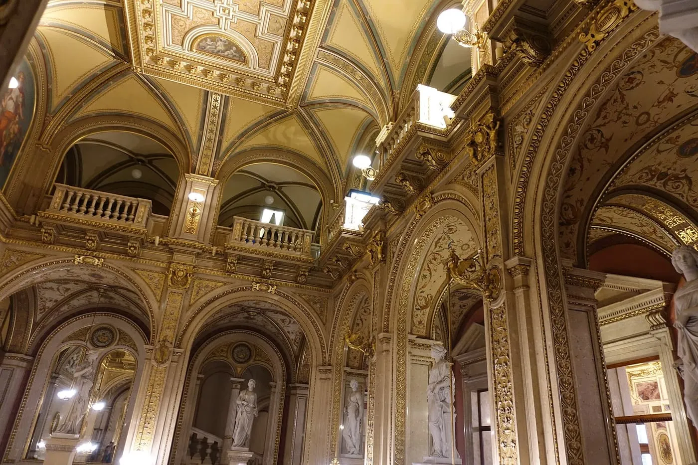 An arcaded hall inside the State Opera with gilded vaults, a balcony and marble statues in arched niches