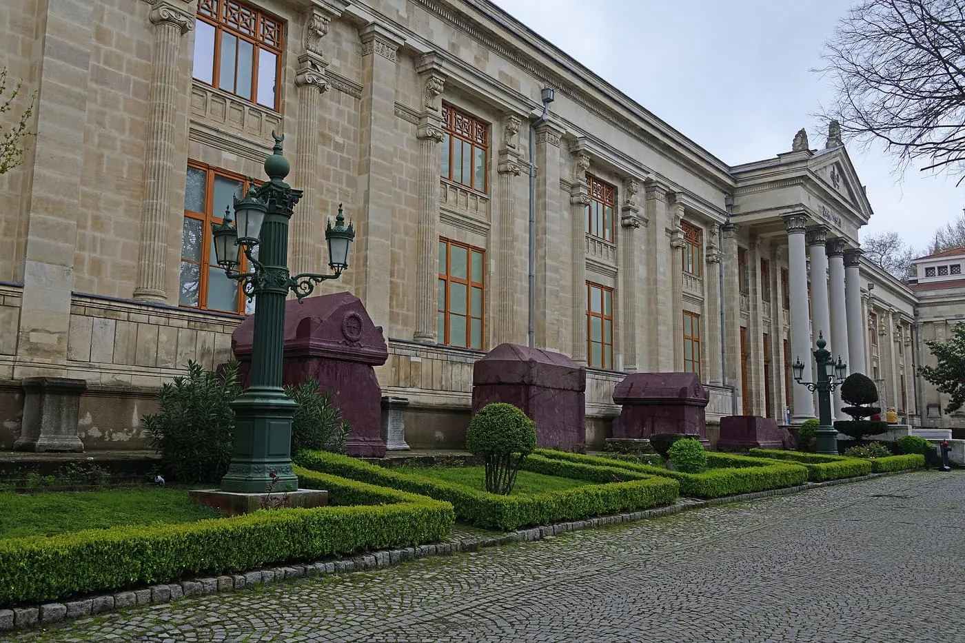 The columned stone facade of the Istanbul Archaeology Museums behind a clipped hedge, with purple-red sarcophagi beside the lawn