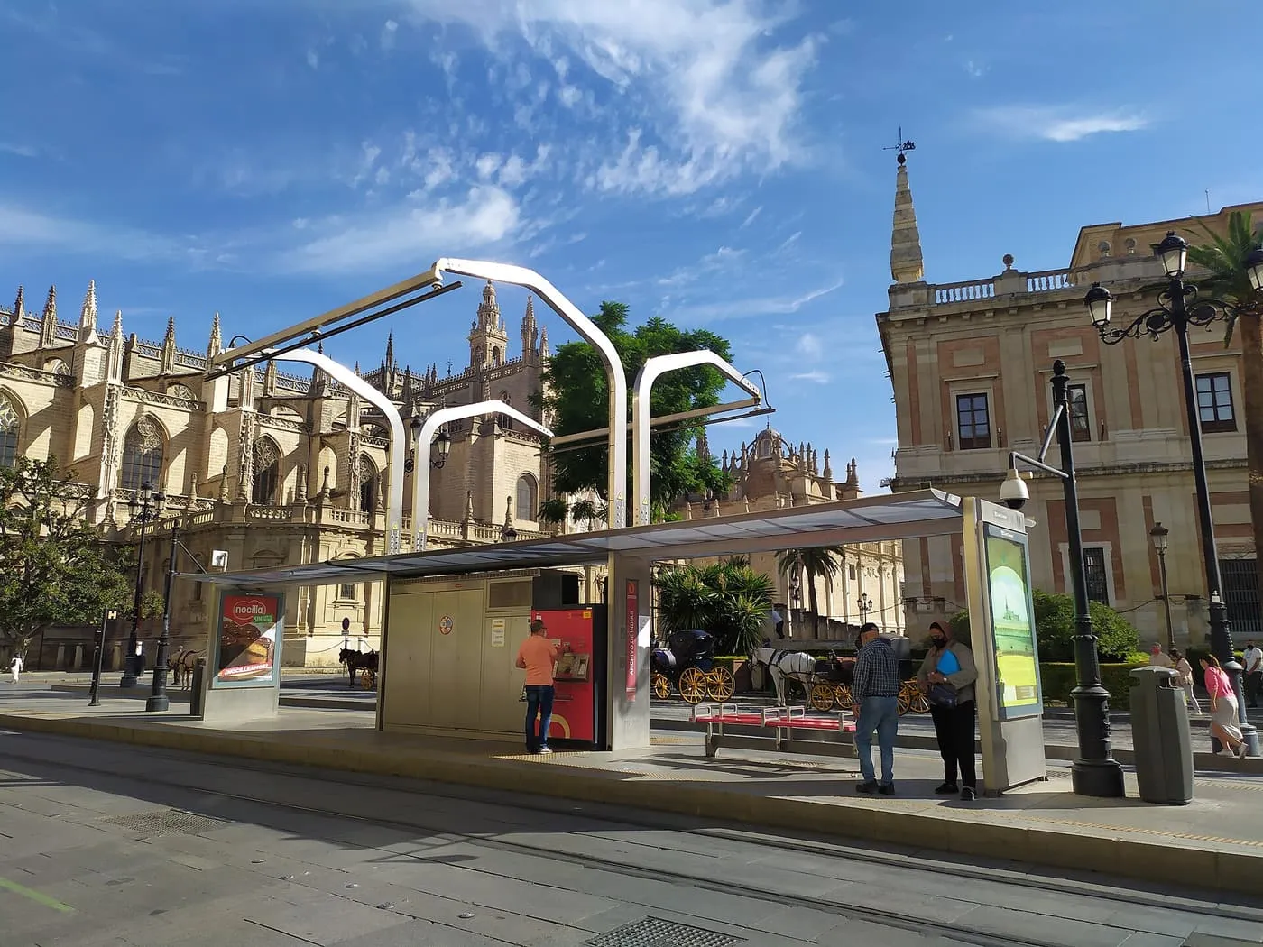 The Metrocentro tram stop at the Archivo de Indias, with a ticket machine and waiting passengers, and the Cathedral and Giralda behind