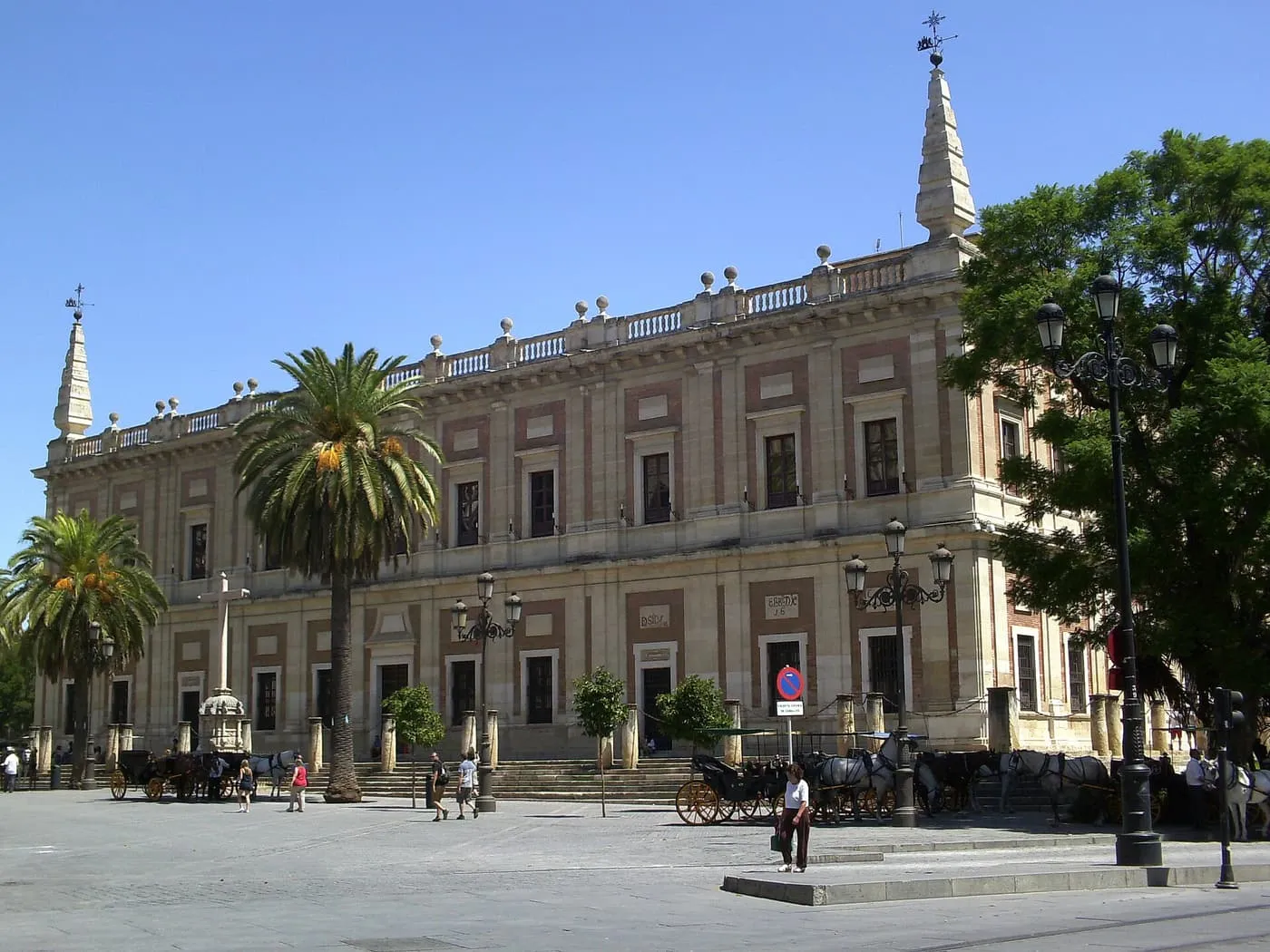 The long stone and brick front of the Archivo General de Indias, with palm trees and horse carriages in front