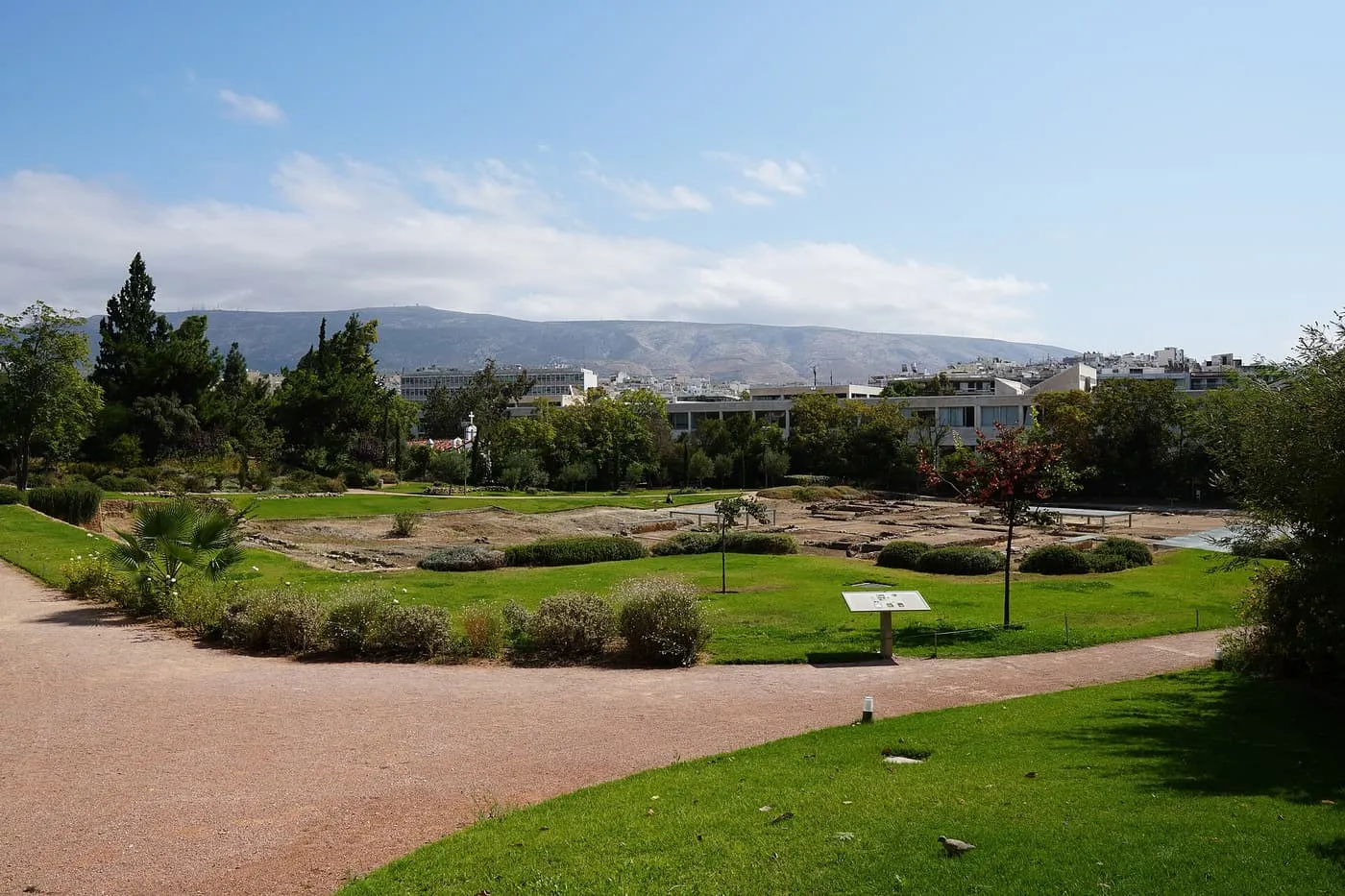 The excavated foundations of Aristotle's Lyceum in a landscaped park in Athens, with lawns, a path and a signboard in front and Mount Hymettus behind