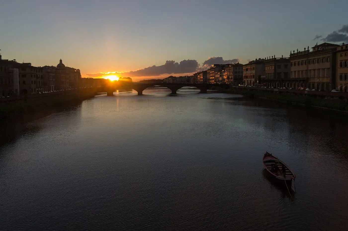 The sun setting over the Arno between two embankments, with a bridge across the river and a rowing boat in the foreground