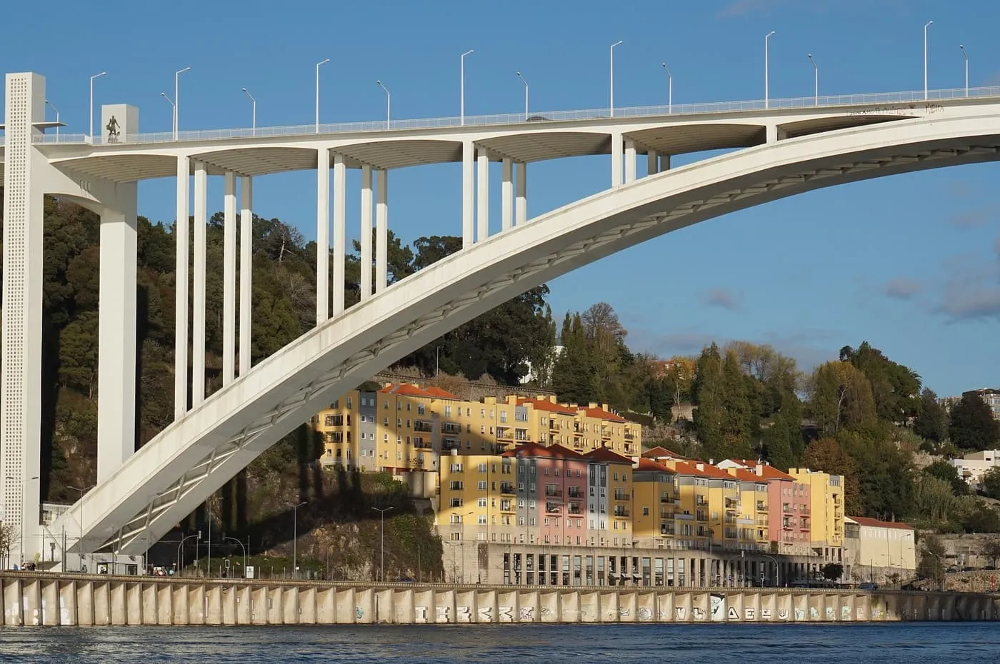 The white concrete arch of the Arrábida bridge seen from the river, with yellow and pink houses on the bank below