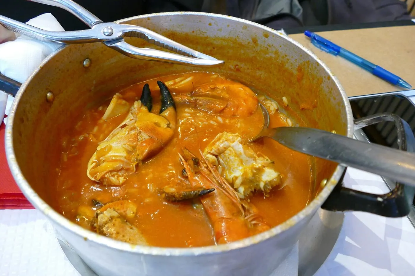 A metal pot of arroz de marisco, tomato-red seafood rice with a crab claw and a prawn, with shellfish crackers on the rim