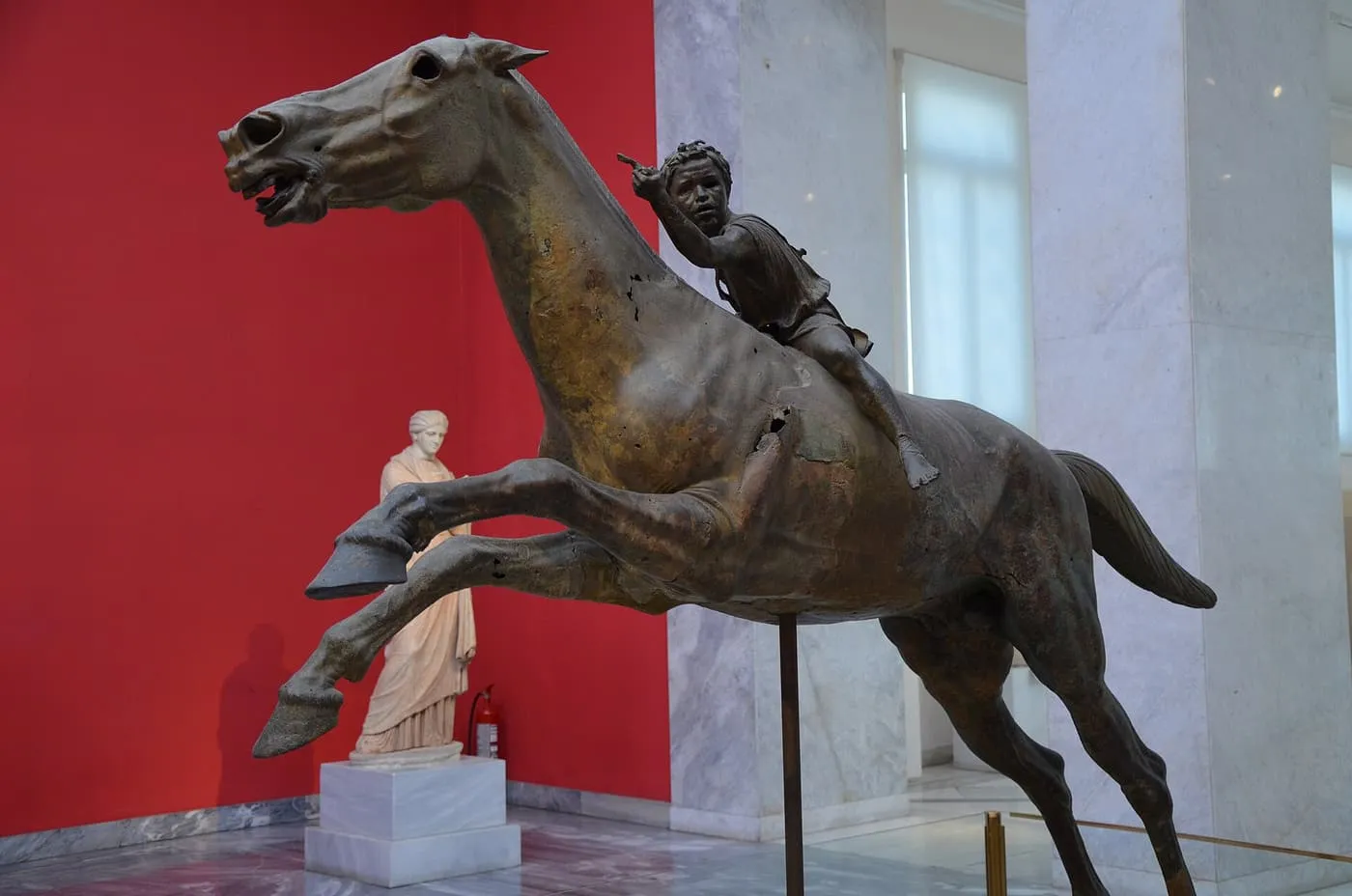 A bronze horse leaping forward with a young jockey on its back, against a red wall