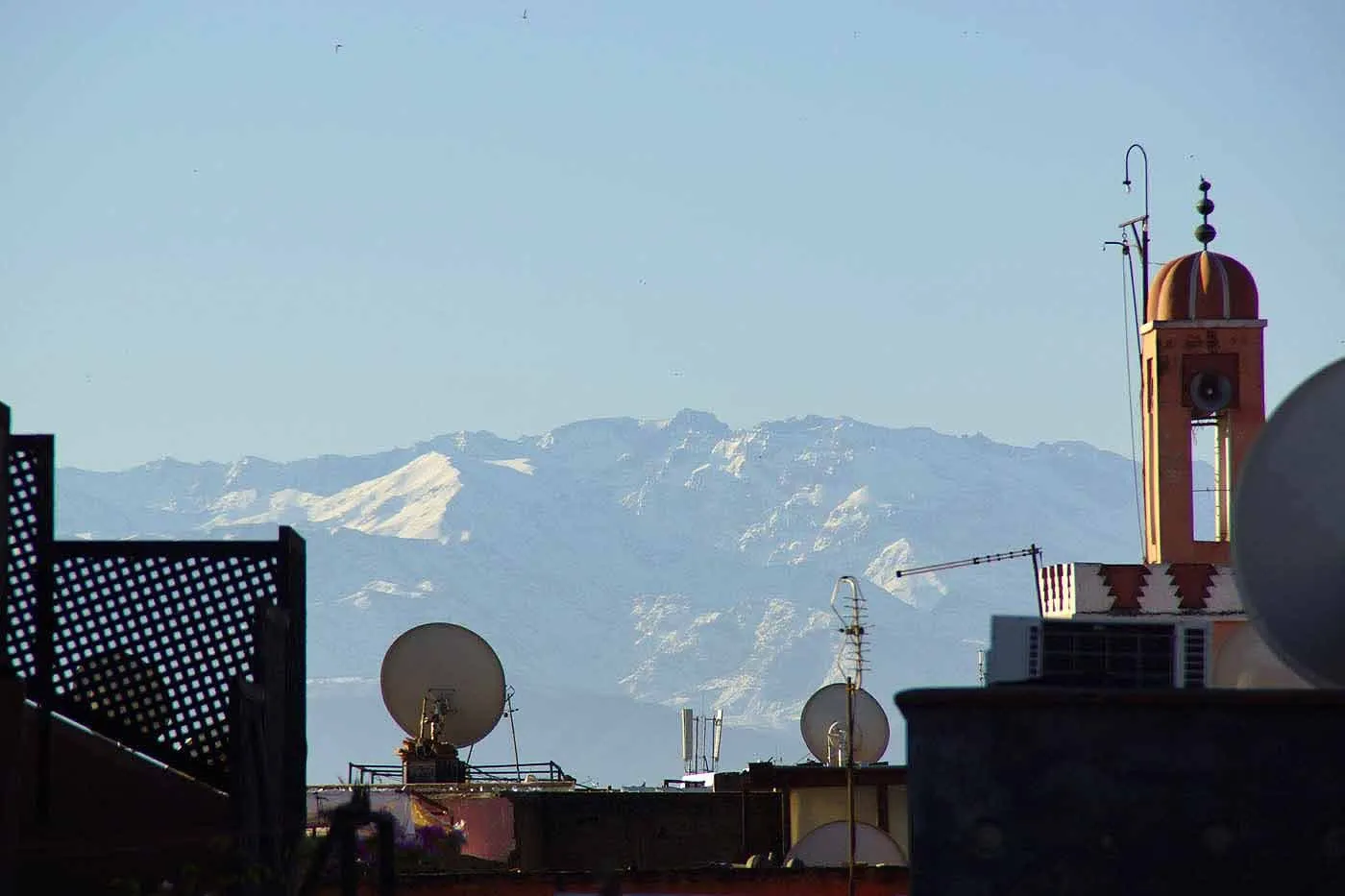 The snow-capped High Atlas on the horizon behind the rooftops of the medina, with a minaret and satellite dishes in the foreground