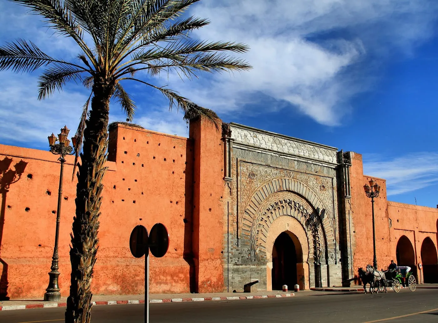 The carved stone gate of Bab Agnaou in a red city wall, with a palm tree in front and a horse-drawn carriage on the road