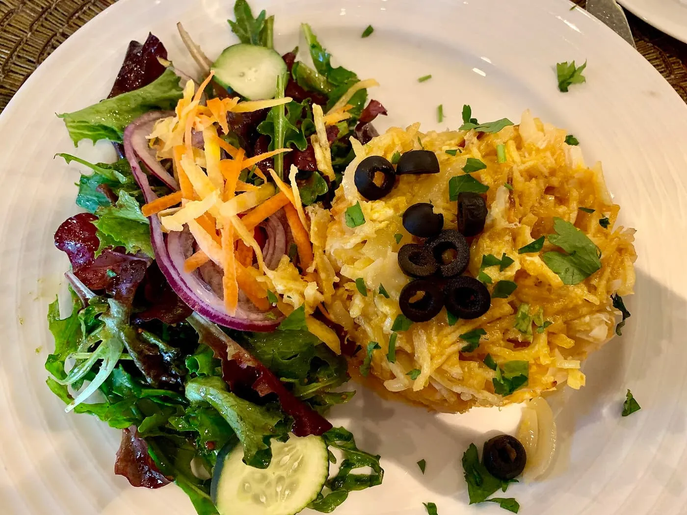 A plate of bacalhau à Brás, shredded salt cod with egg and fried potato, topped with black olives and parsley, beside a green salad