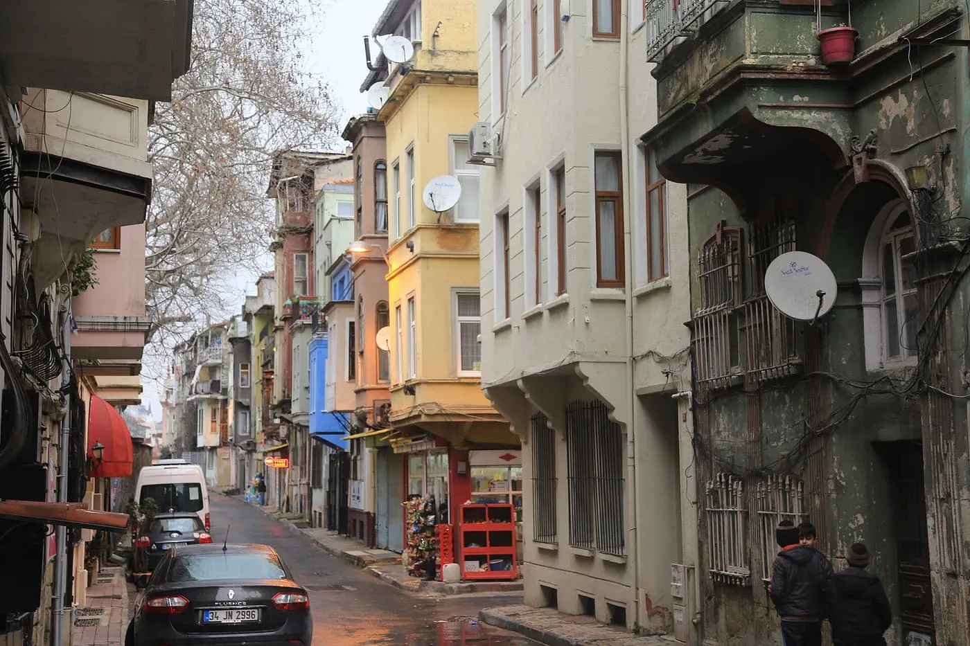A sloping Balat street of tall, narrow houses in yellow, blue, green and cream, with a parked car in the foreground and a bare plane tree overhead