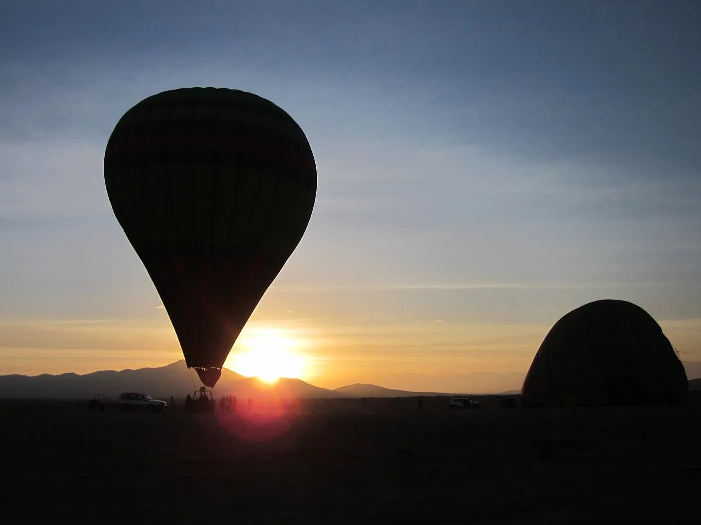 A hot air balloon silhouetted against the low sun with hills on the horizon, and a second balloon laid out on the ground to the right