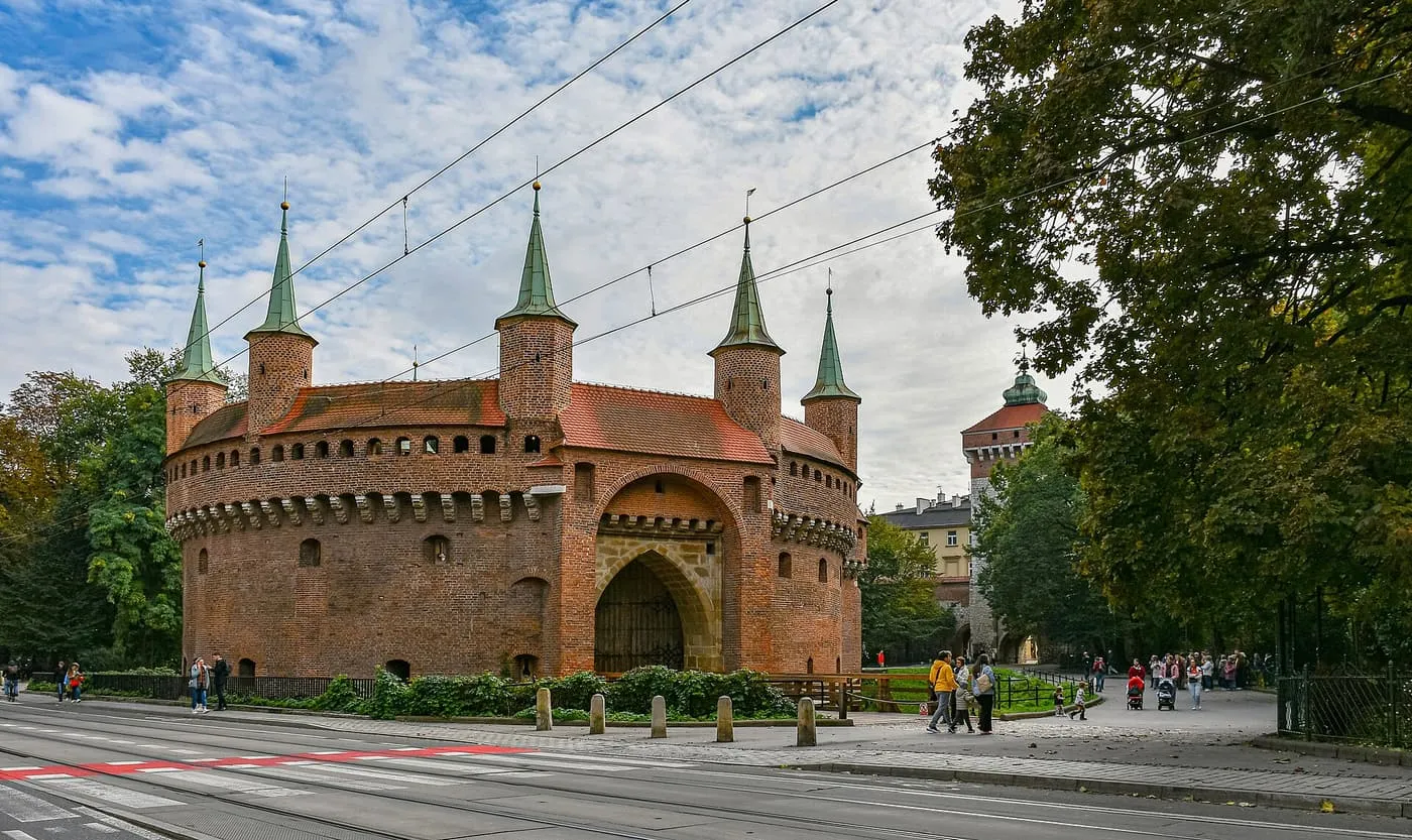 The Barbican, a round red-brick fortress with five green-roofed turrets, seen across Basztowa Street, with the tower of St Florian's Gate behind trees on the right