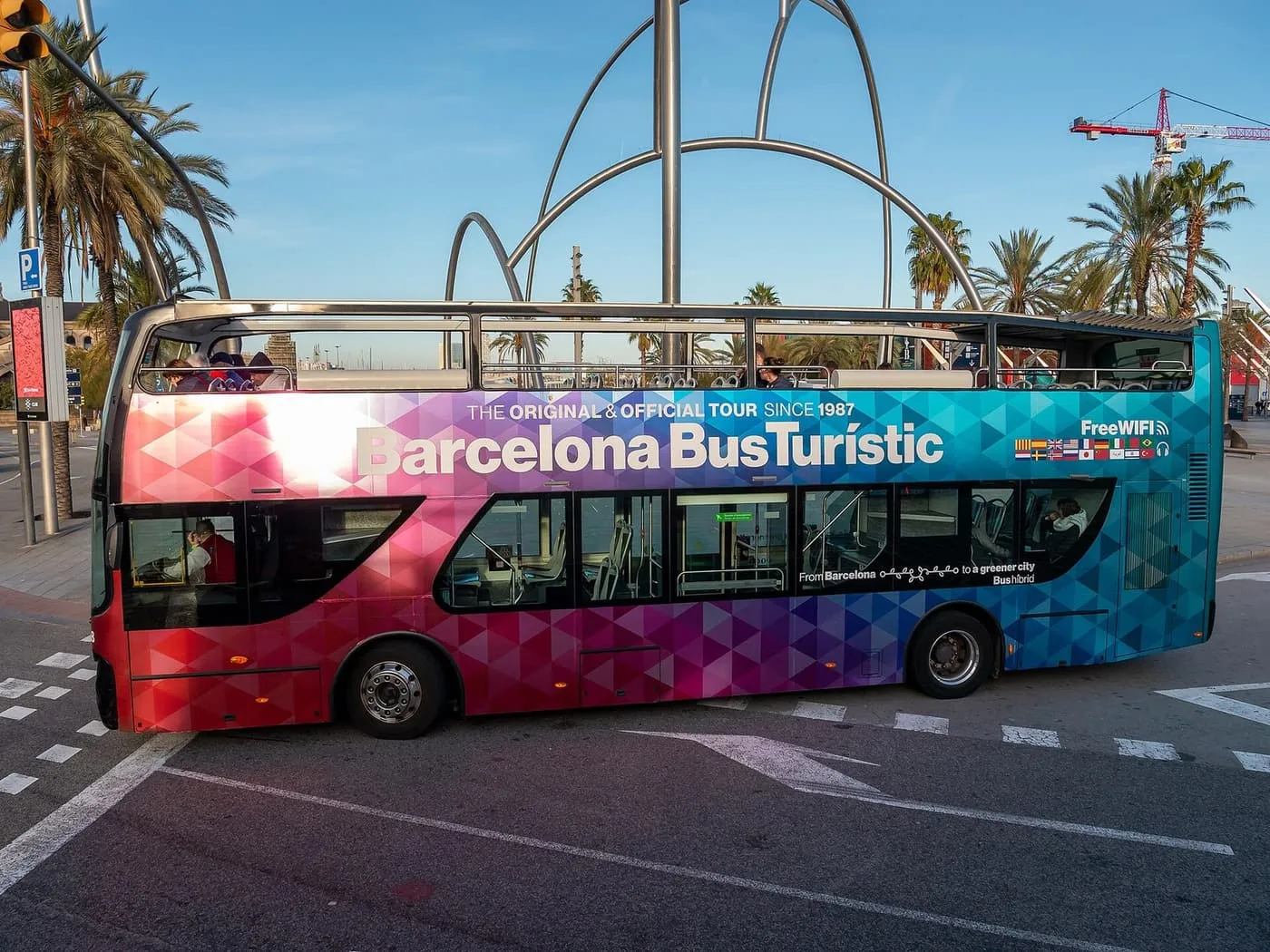 A double-decker open-top Barcelona Bus Turístic in a colourful geometric livery turning a corner, with palm trees behind