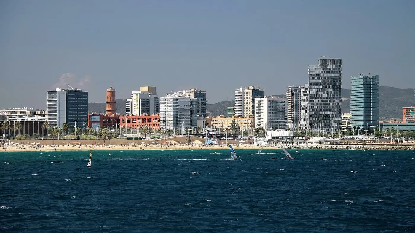 Barcelona's seafront from the sea: a sandy beach with windsurfers off it and a row of high-rise blocks and a brick tower behind