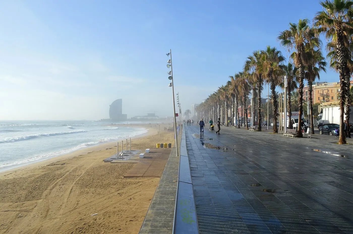 Barceloneta beach with waves on the sand, a wide promenade lined with palms, and a tall glass tower far off along the shore