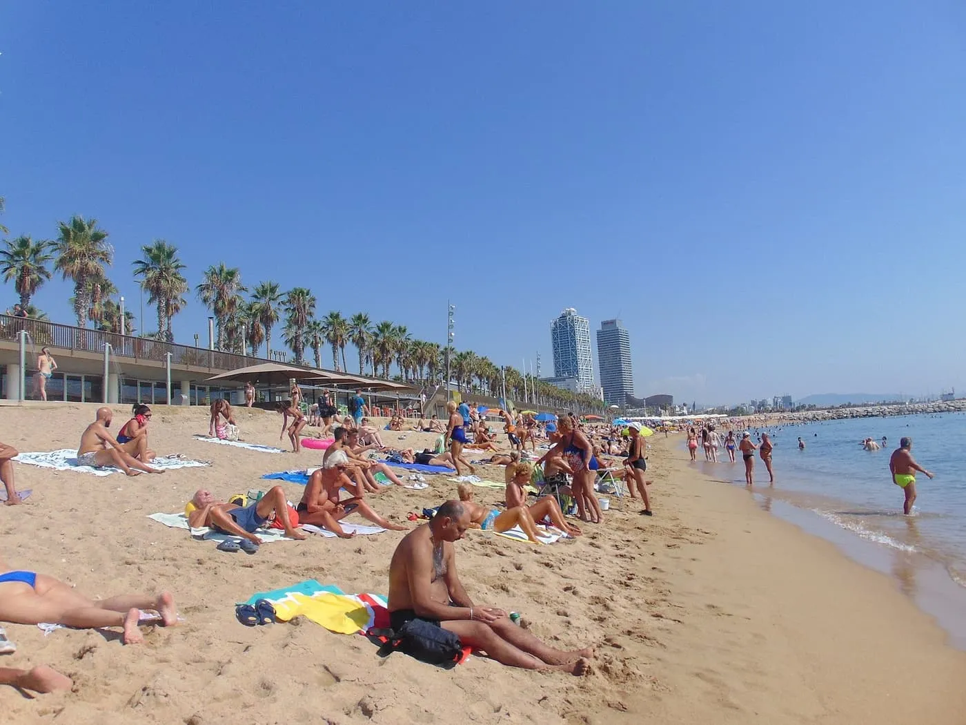Sunbathers on the sand of Barceloneta beach, with the promenade and its palms on the left and the towers of the Port Olímpic in the distance