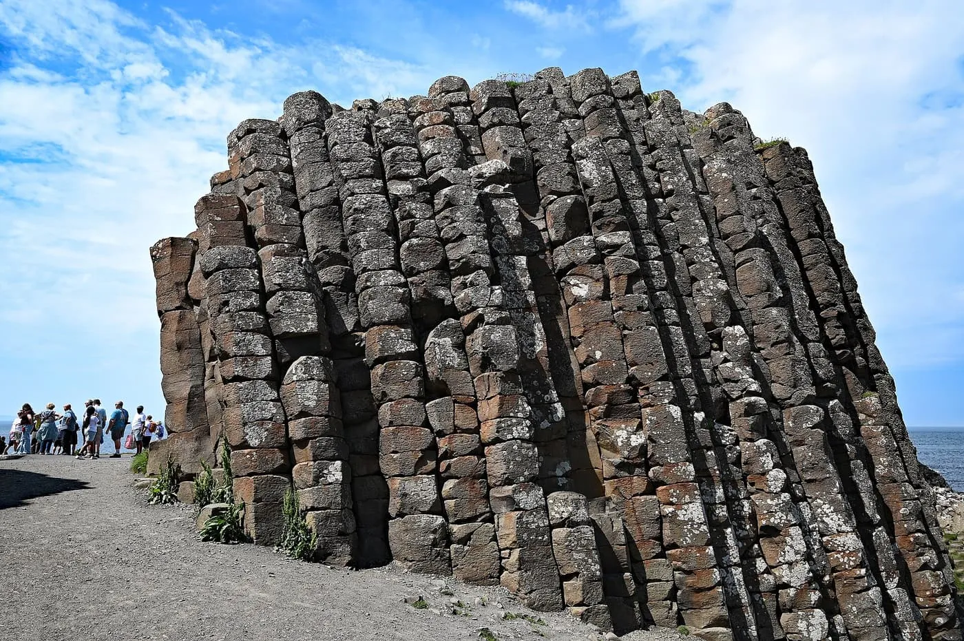A tall block of vertical basalt columns beside a gravel path, with a small group of visitors beside it