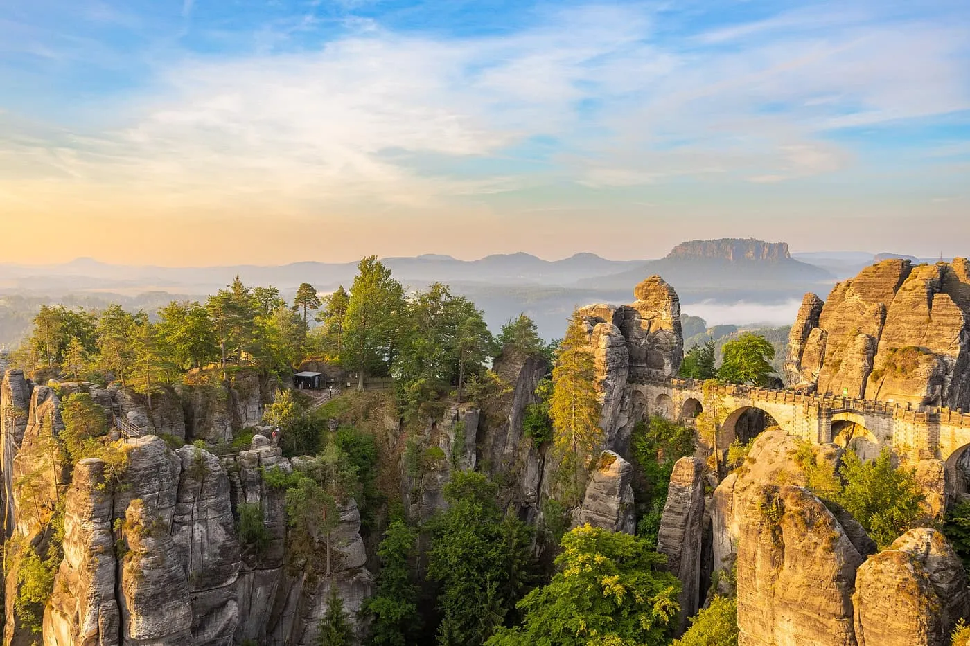 The stone Bastei Bridge crossing between sandstone pillars above forested valleys in low golden light, with a flat-topped hill on the horizon