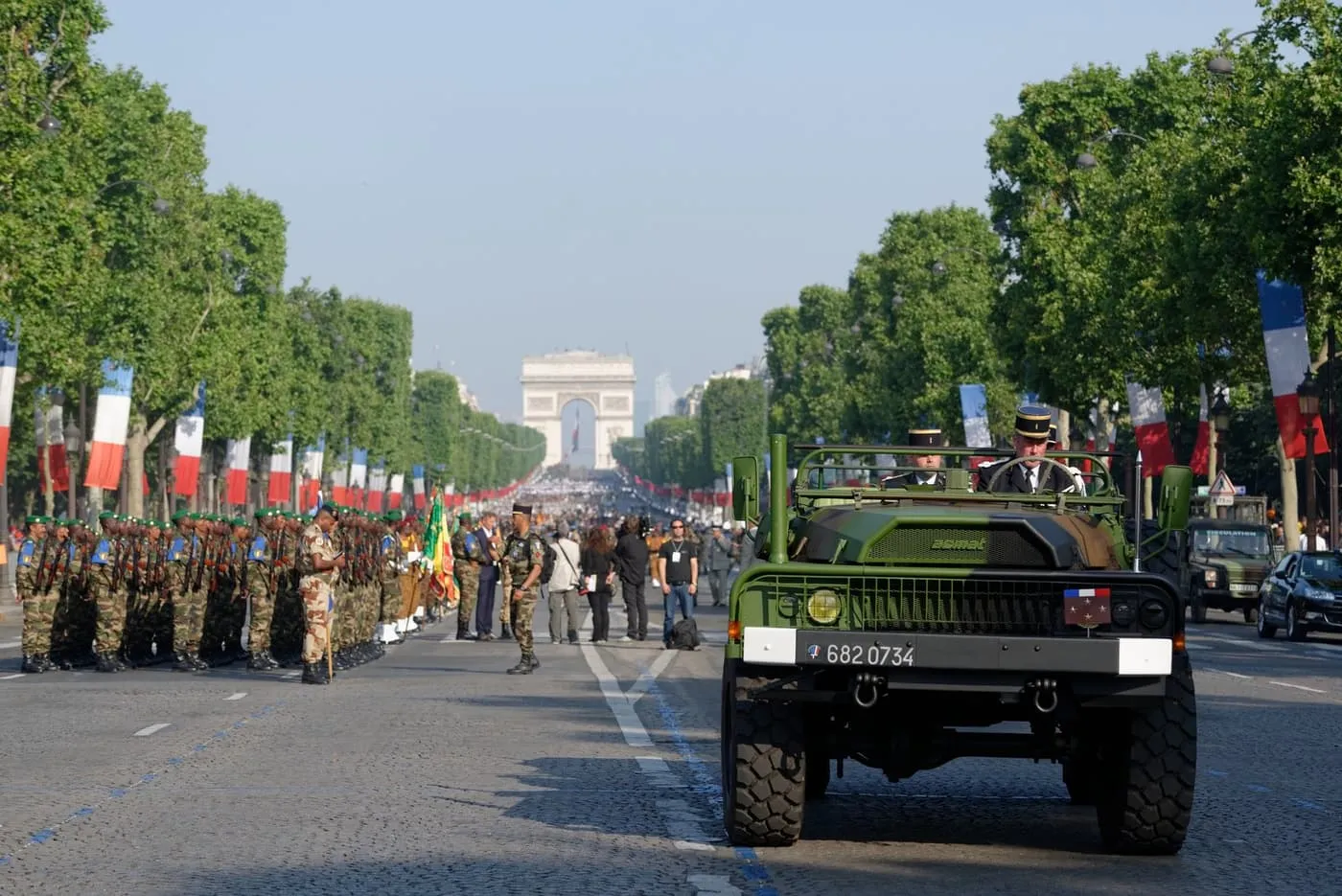 Soldiers in camouflage lined up on the Champs-Élysées beside a military jeep, between rows of tricolour banners, with the Arc de Triomphe at the far end